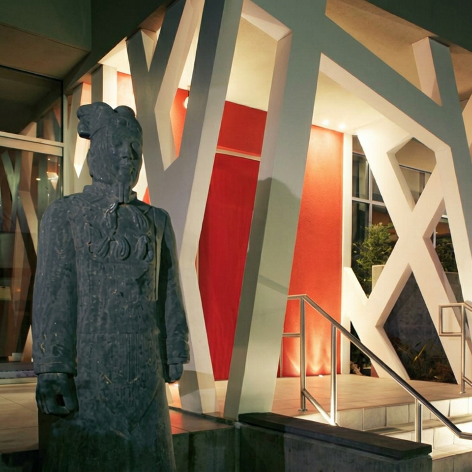 Nighttime restaurant entrance with a dark statue, dramatic white geometric columns, red walls, glass doors, and illuminated stairs.