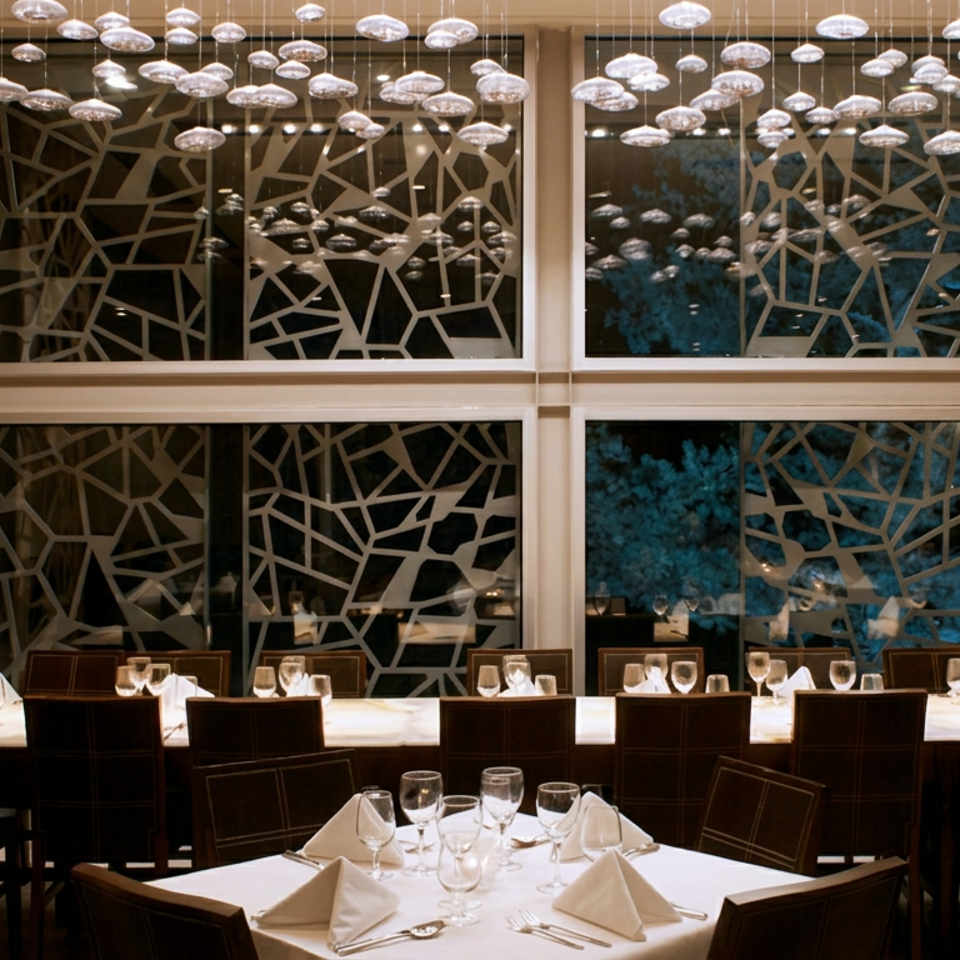 Elegant restaurant dining room with white tablecloths, folded napkins, glassware, geometric window screens, and suspended pendant lights.