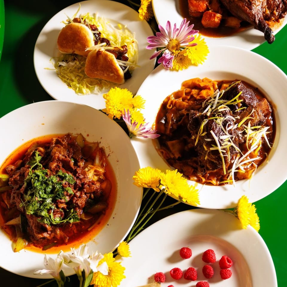 Overhead spread of richly sauced dishes, stuffed buns, flowers, and raspberries arranged on white plates against a green background.