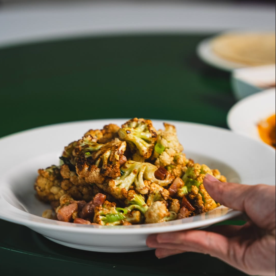 Roasted cauliflower florets with green herbs and browned vegetables served on a white plate, held over a dark green table.