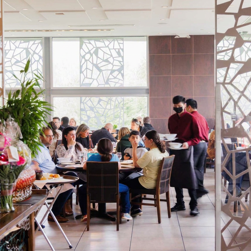 Busy restaurant dining room with seated guests, red-uniformed servers, bright windows, plants, and decorative geometric partitions.