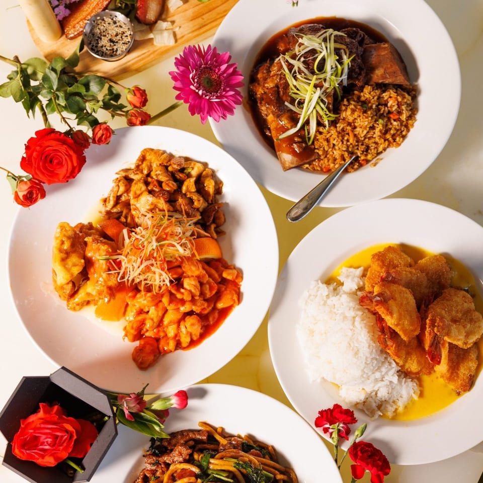 Overhead spread of colorful Asian-inspired dishes with rice, sauced entrées, vegetables, and decorative roses on white plates.
