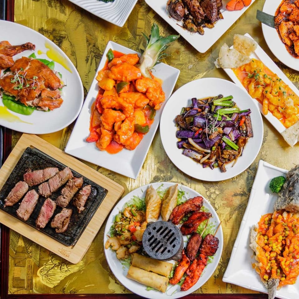 Overhead spread of colorful Asian dishes, including roasted chicken, orange-glazed seafood, grilled steak, vegetables, spring rolls, and fish.