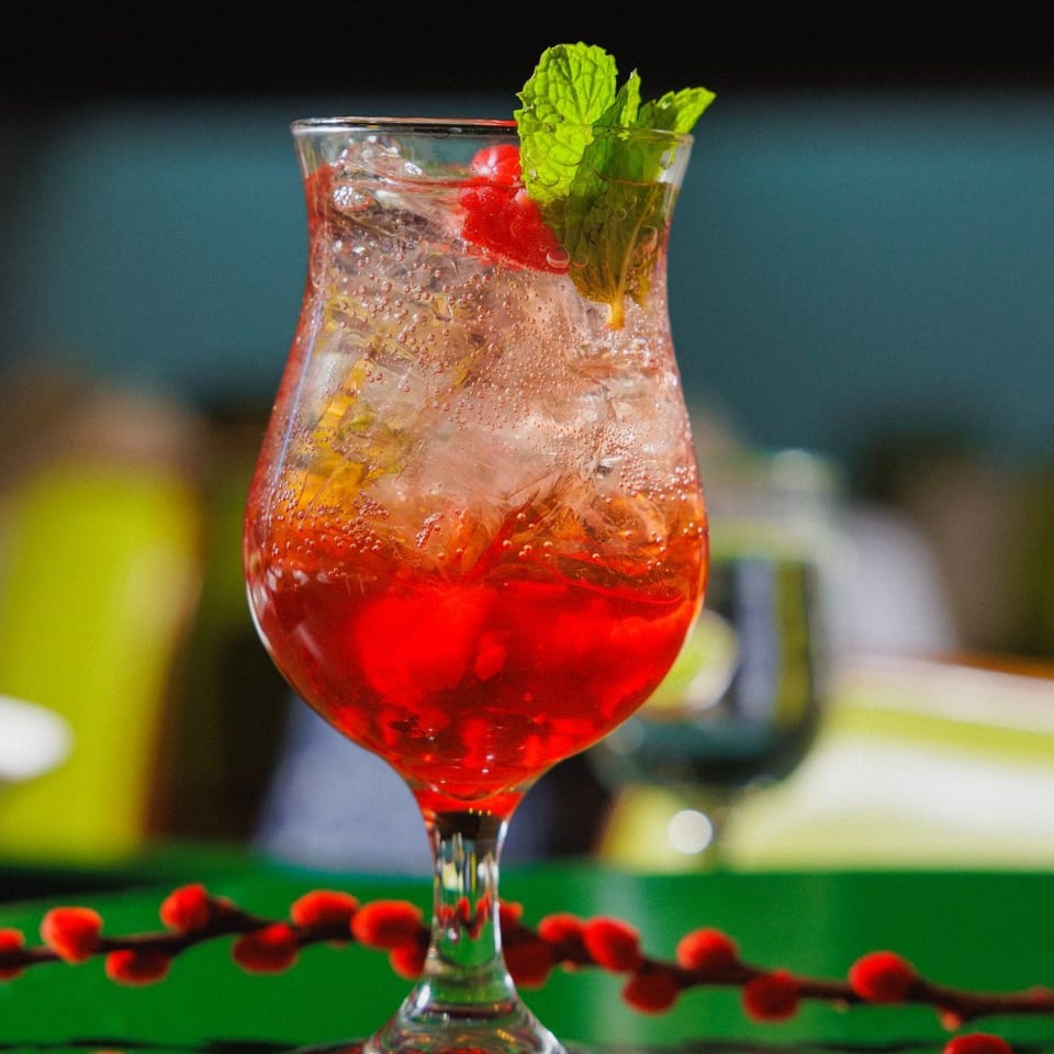 Red fruit cocktail with ice, mint leaves, and berries served in a stemmed glass against a blurred restaurant background.